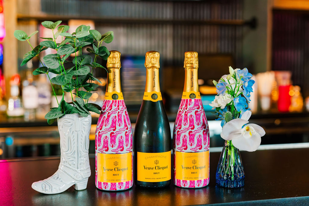 Three customized bottles of champagne with pink boots on the label sitting between a statue