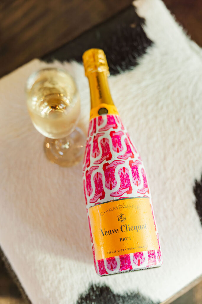 Customized pink boots champagne bottle laying next to a glass of champagne on top of a cow patterned stool