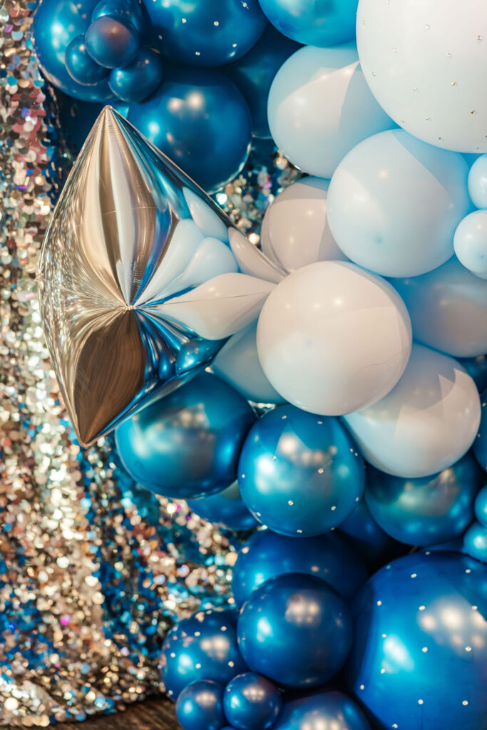 Blue and white balloon garland draping from the top right of a silver glitter backdrop