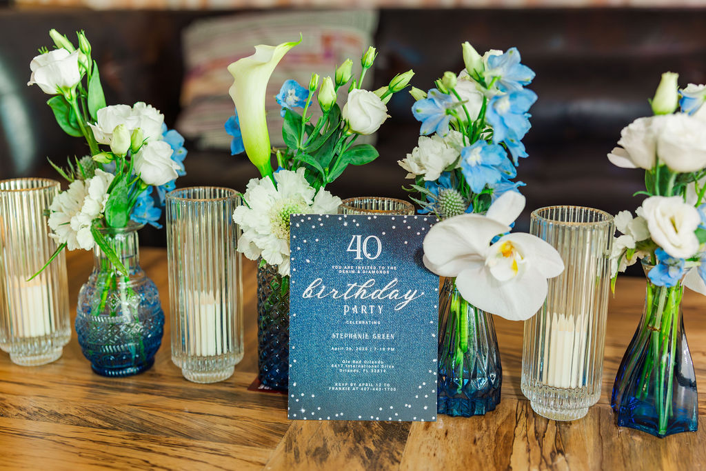 40th Birthday stationary sitting in front of blue and white floral centerpieces with tall white votives for candles