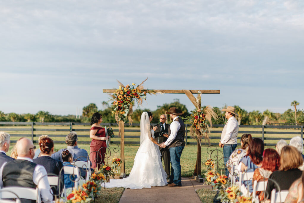 Wide-angle view of an outdoor country wedding ceremony with guests seated, and a bride and groom standing under a decorated wooden arch.