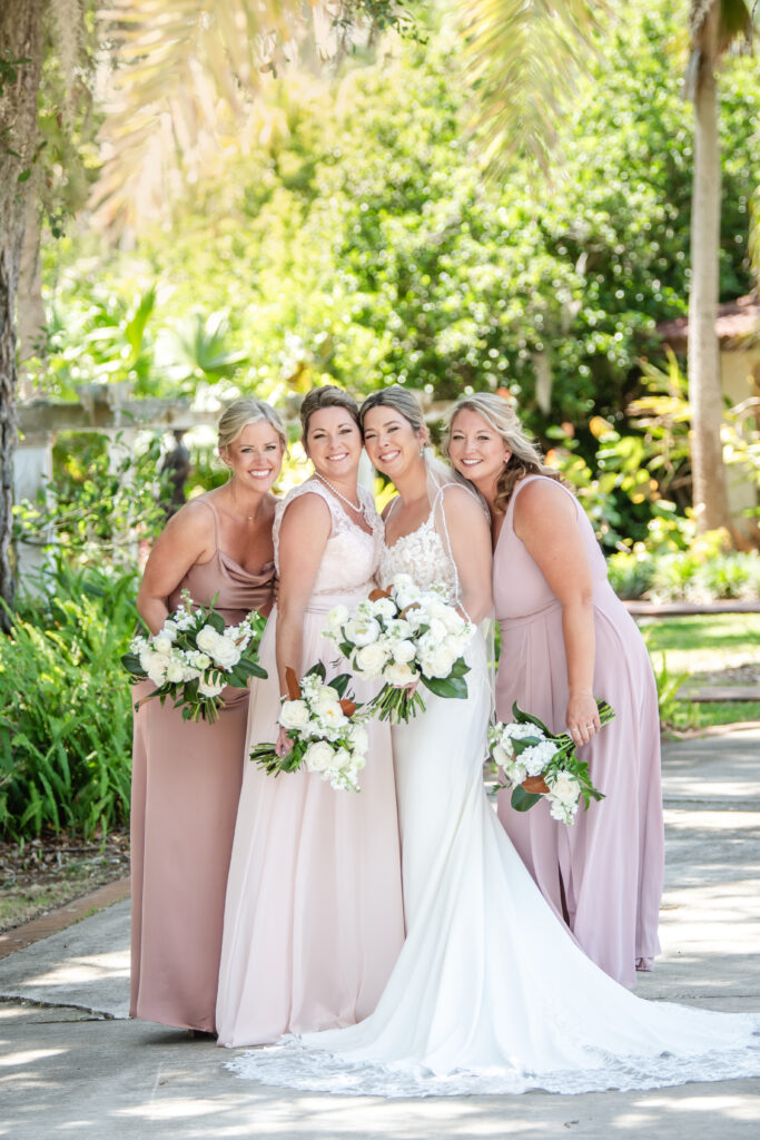 Bride posing with bridesmaids in blush and dusty rose dresses, all holding white floral bouquets in a sunny garden setting.