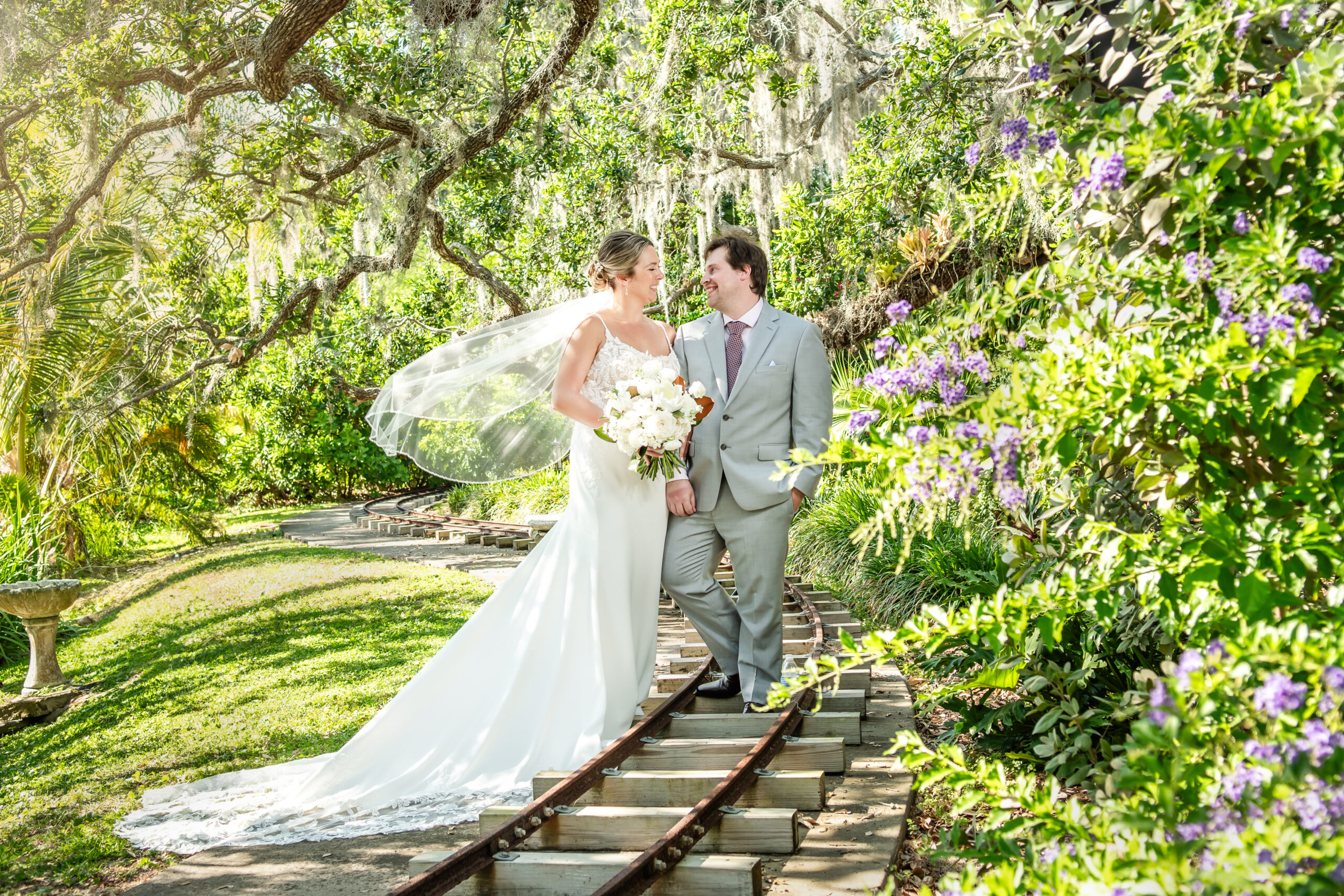 Bride and groom standing together on garden train tracks, smiling at each other under lush greenery with the bride’s veil flowing.