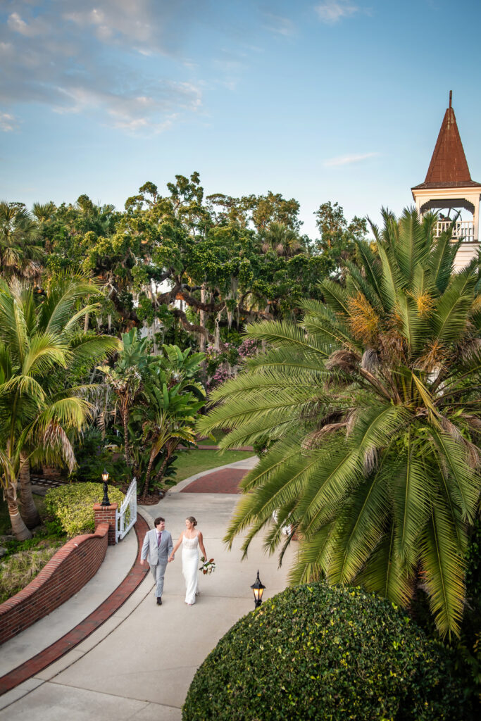 Bride and groom walking hand-in-hand along a curved pathway surrounded by tropical greenery and palm trees at a garden venue.