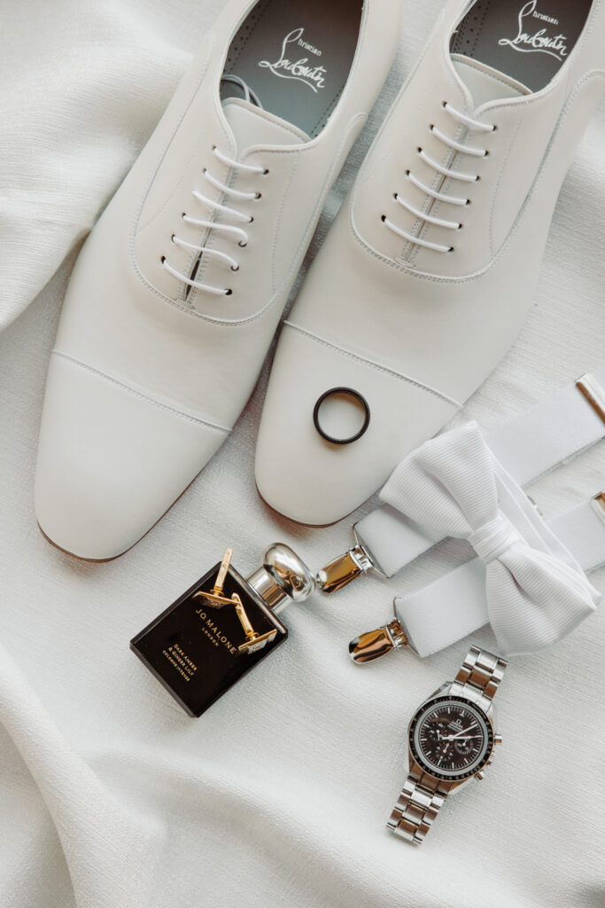 Flat lay of groom's wedding accessories including white Christian Louboutin shoes, a black ring, a white bow tie, cufflinks, a watch, and cologne.