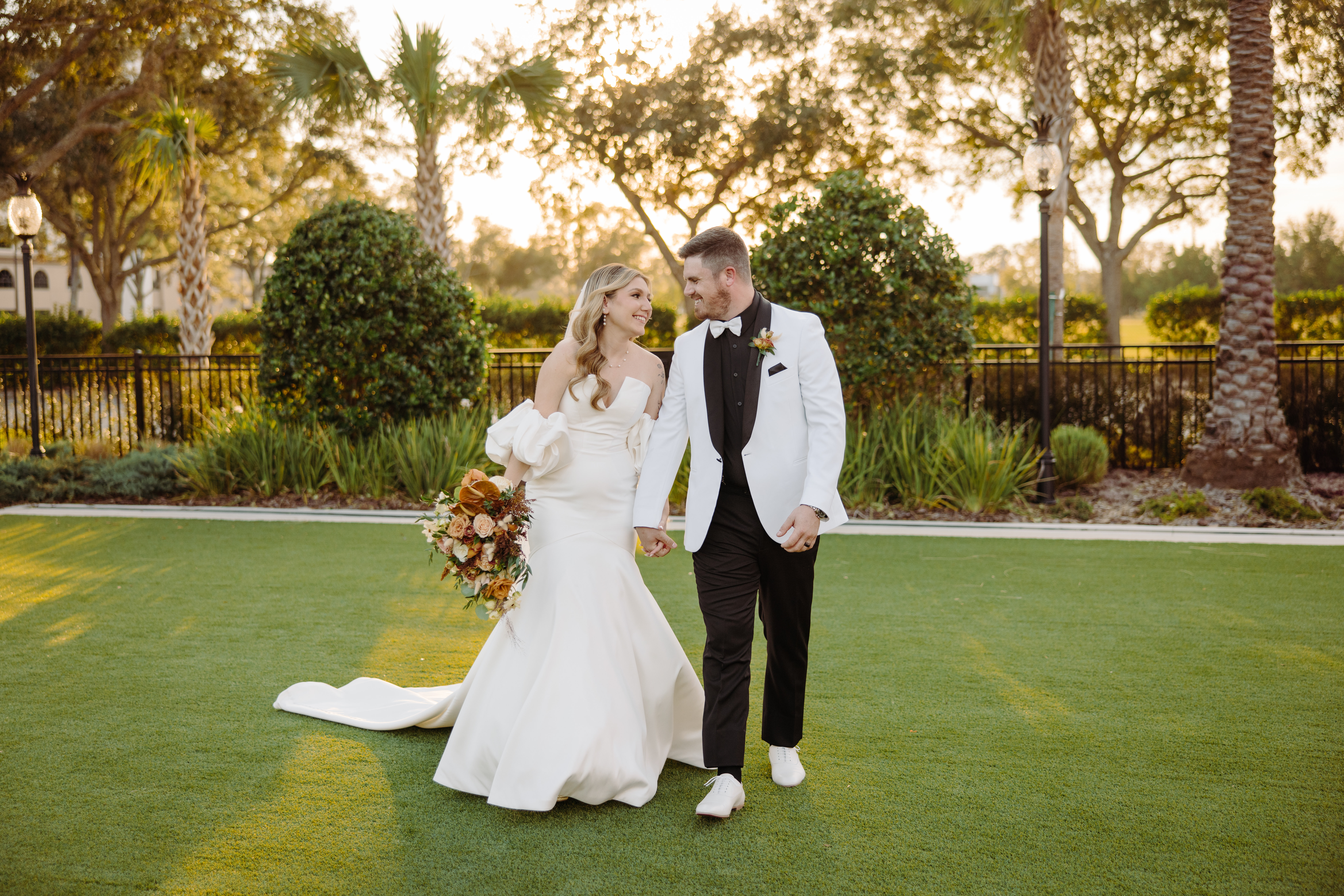 Bride and groom walking hand in hand across a lush lawn surrounded by palm trees, sharing a warm look at sunset.