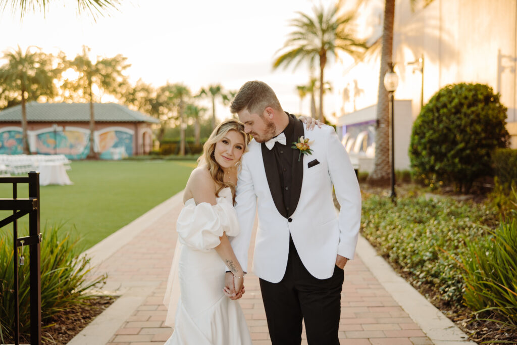 A bride in an off-the-shoulder gown and a groom in a white tuxedo jacket walk together outdoors at sunset, smiling and holding hands among palm trees.