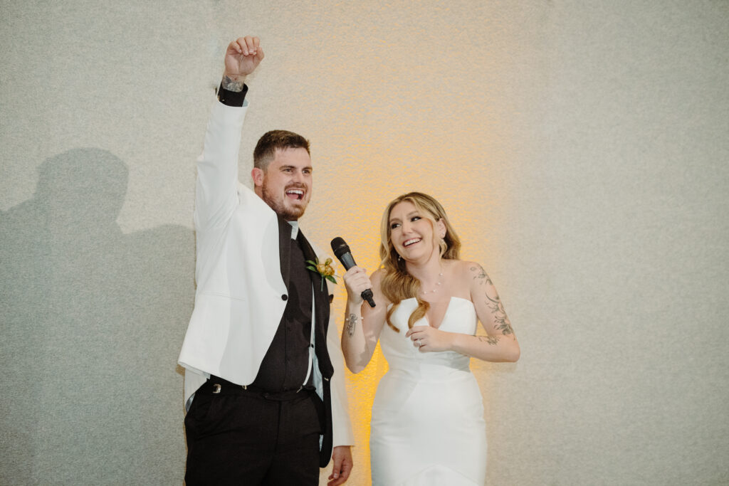 Bride and groom laughing during their wedding reception, the groom raising his arm triumphantly while the bride holds a microphone