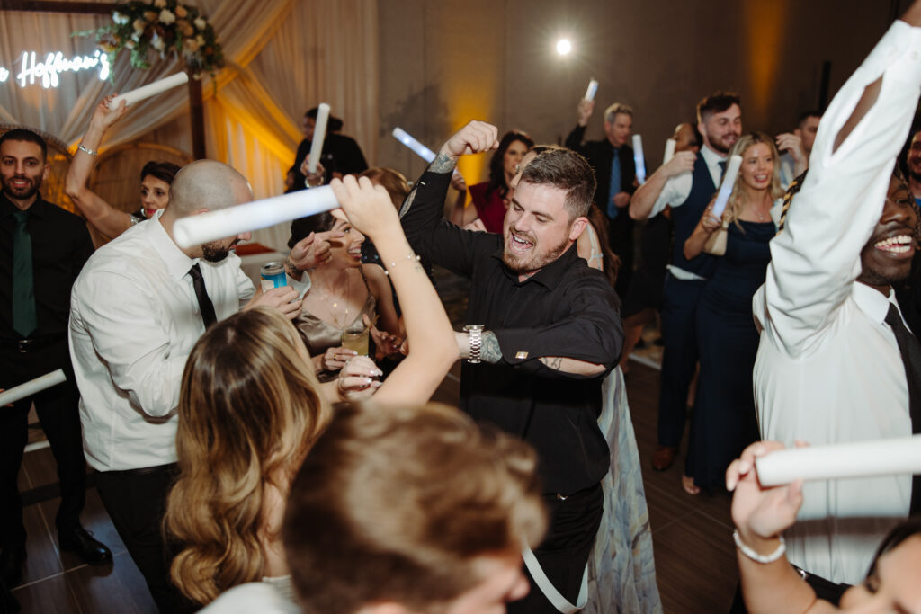 Groom in black shirt dancing joyfully among wedding guests holding glow sticks during a lively reception.