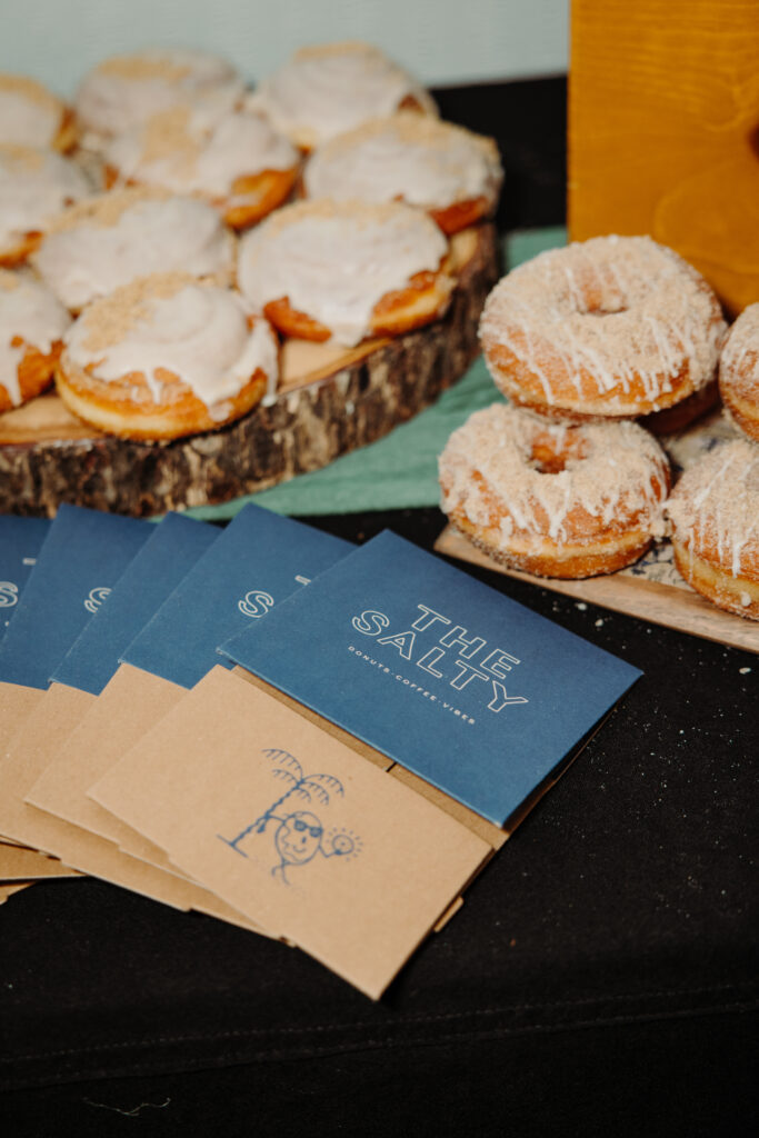 Wedding dessert table featuring gourmet donuts from The Salty, displayed on rustic wood stands with branded favor cards