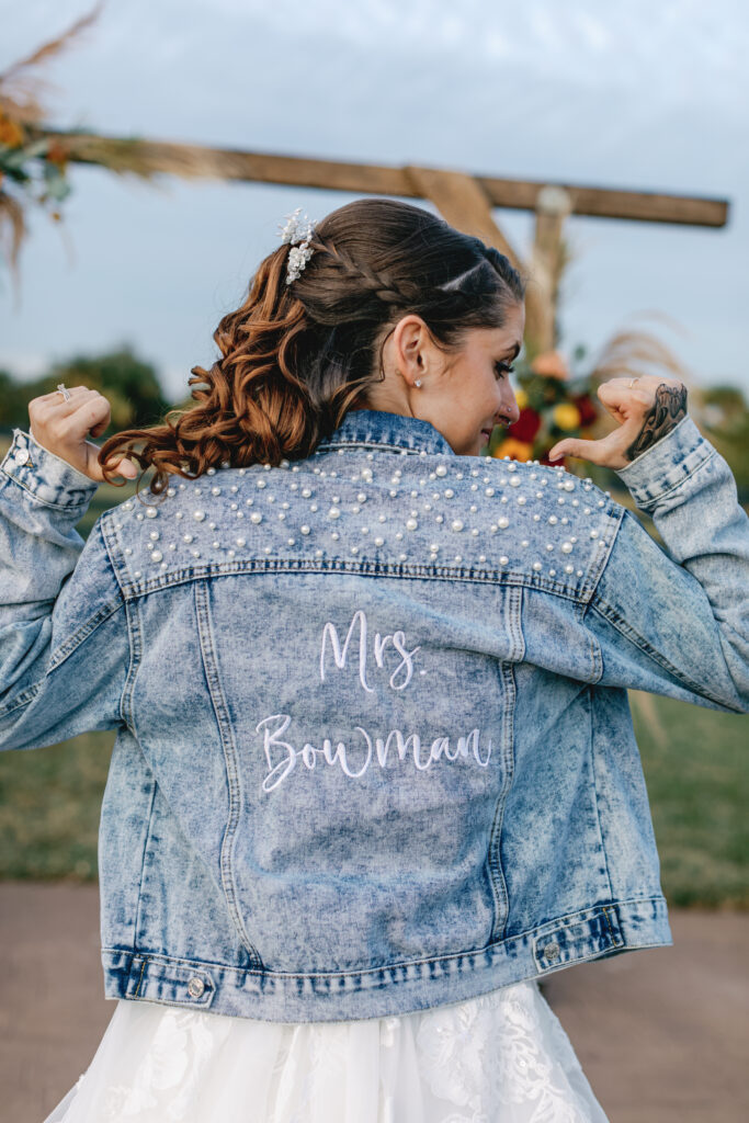 Bride wearing a custom pearl-studded denim jacket embroidered with "Mrs. Bowman" over her wedding dress