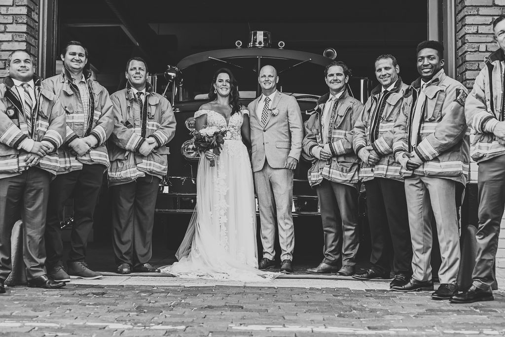 Bride and groom pose in front of a fire truck with fireman in uniform, creating a unique wedding photo