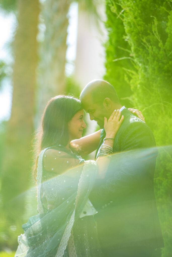 Romantic outdoor portrait of a bride and groom in traditional Indian attire holding each other forehead to forehead surrounded by greenery