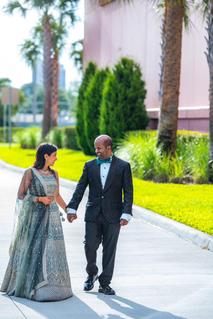 A bride and groom holding hands and walking outdoors, dressed in formal wedding attire, smiling at each other.
