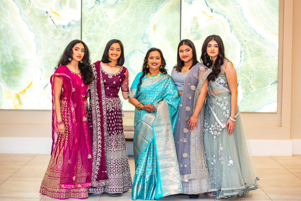 Group portrait of women in colorful traditional Indian attire, including lehengas and a saree, smiling and posing together in front of a modern marble-style backdrop.