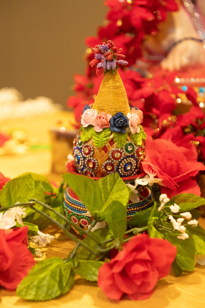 Close-up of a decorative kalash adorned with colorful beads, flowers, and fabric surrounded by red roses.