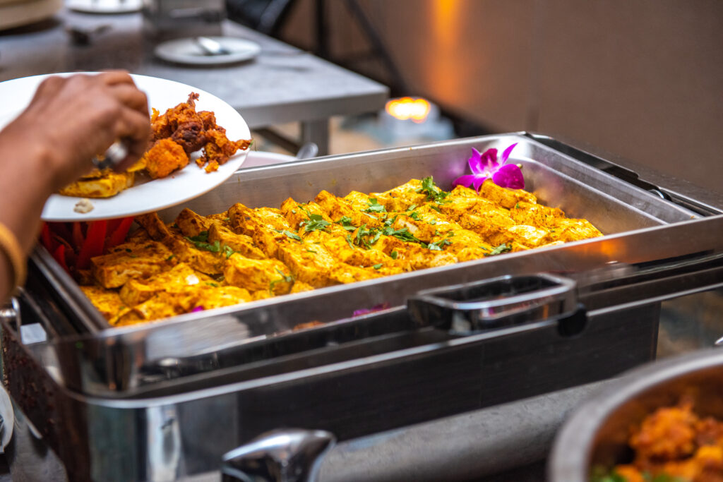 Tray of golden paneer tikka garnished with fresh herbs, served in a buffet with a guest filling their plate.