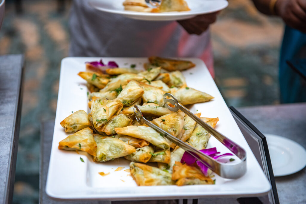 White platter with crispy triangular samosas garnished with herbs, displayed at a wedding buffet with serving tongs.
